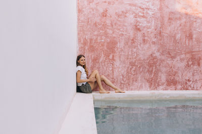 Full length portrait of a blond, caucasian woman sitting against a colored wall by the poolside