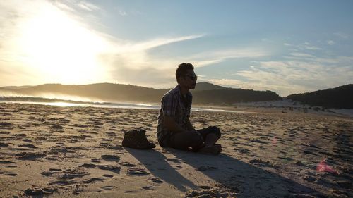 Silhouette young man sitting at beach against sky on sunny day