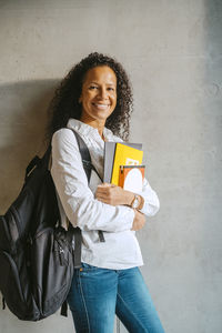 Portrait of smiling woman holding book and backpack leaning on gray wall at college