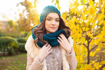 Portrait of beautiful young woman in warm clothing standing against autumn tree