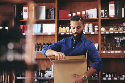 Salesman unpacking cardboard box while standing against rack in deli