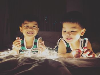 Smiling brothers with illuminated string lights lying on bed in darkroom at home