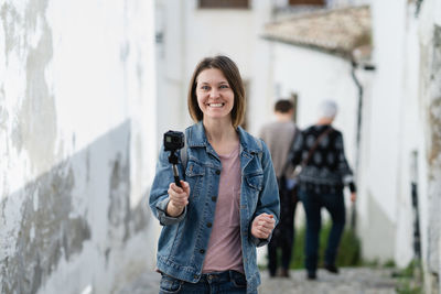 Portrait of smiling young woman holding camera while standing in alley