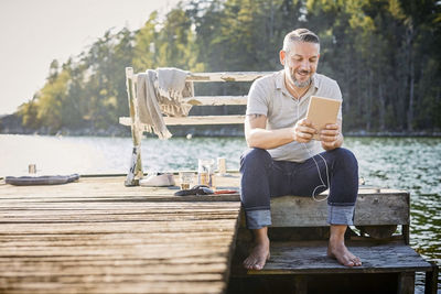 Smiling mature man using digital tablet with in-ear headphones while sitting on jetty over lake