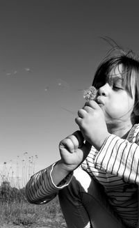 Girl blowing dandelion seed while crouching against clear sky
