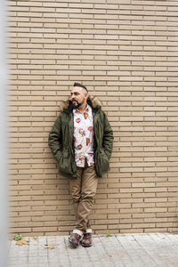 Young bearded man with hands on pocket leaning on wall, looking camera