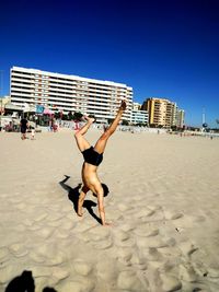 Full length of shirtless man doing handstand on sand at beach against blue sky
