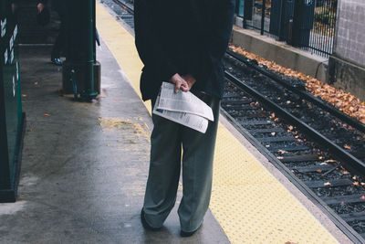 Low section of man holding newspaper while standing at railroad station platform