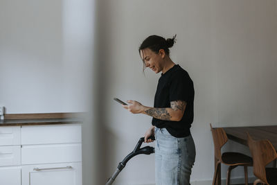 Smiling woman using cell phone while vacuum cleaning room