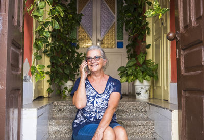 Cheerful senior woman talking on mobile phone while sitting on steps at home