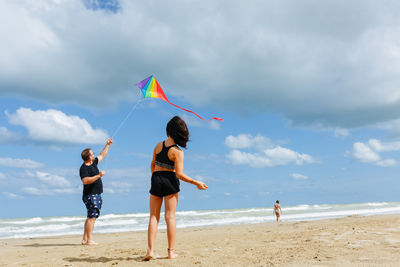 Rear view of woman with arms outstretched standing at beach against sky