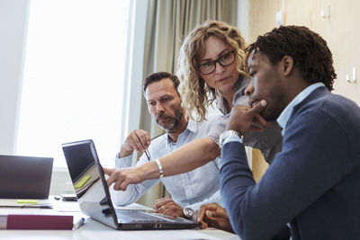 Serious mature businesswoman looking at male colleague while using laptop in board room