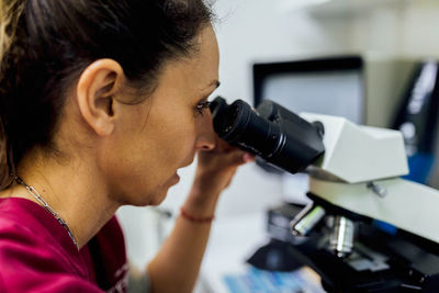 Side view of focused female veterinarian in uniform looking through microscope while checking results of medical procedure in modern clinic