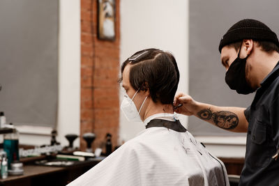 Barber in protective mask and latex glove styling adult man in a barbershop salon. new normal