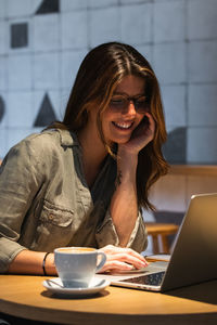 Young cheerful female browsing internet on portable computer at table with hot drink in cafeteria