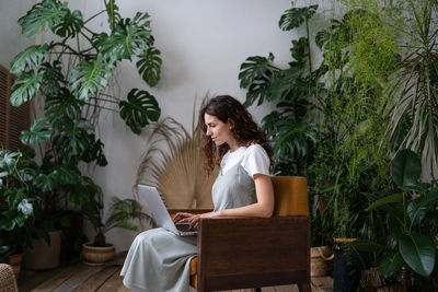 Young woman florist houseplants shop owner working on laptop at home at indoor garden in internet