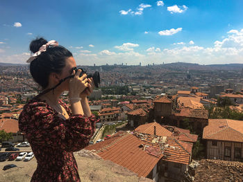Young woman photographing while standing by buildings in city against sky