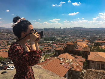 Young woman photographing while standing by buildings in city against sky