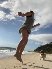 Low angle view of woman jumping by dog at beach against sky