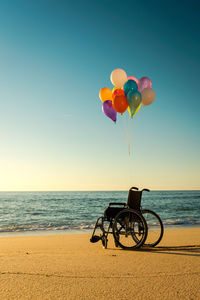 Silhouette wheelchair with colorful balloons on beach against clear blue sky during sunset