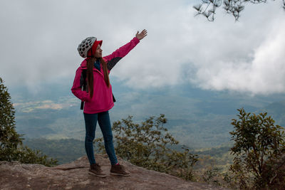 Full length of young woman with hand raised standing on mountain against cloudy sky