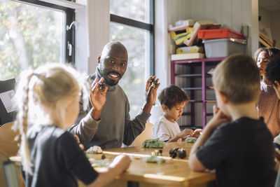 Smiling male teacher holding clay while sitting with kids in classroom at preschool