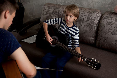 Young male musician teaches little boy student how to play the acoustic guitar and play by notes.
