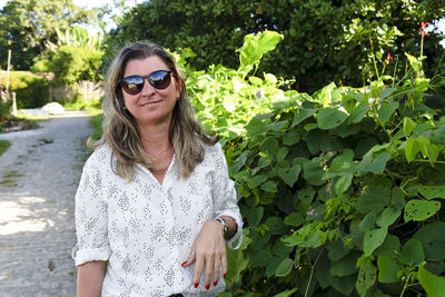 Portrait of a woman wearing sunglasses on a street with plants and green leaves in the background. 