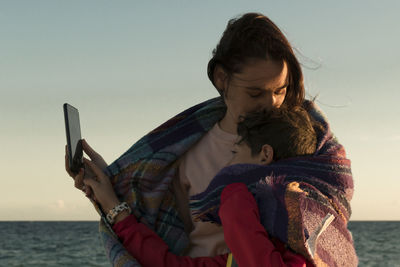 Mother embracing son with smart phone against sea and sky during sunset