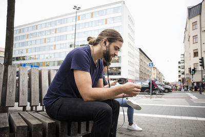 Man listening music through smart phone while sitting beside woman on wooden bench in city
