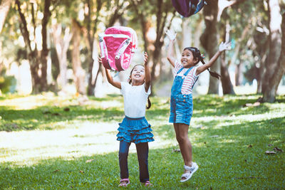 Full length of schoolgirls throwing backpacks in park