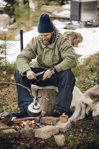 Smiling man wearing warm clothing looking at tea kettle at bonfire