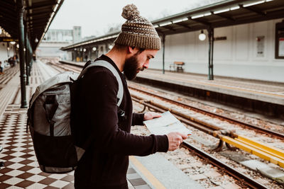Side view of man reading map while standing at railroad station