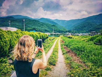 Rear view of woman photographing from mobile phone by agricultural field on dirt road