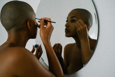 Side view of african american female with bald head doing makeup and applying liquid eyeliner with brush while looking in mirror