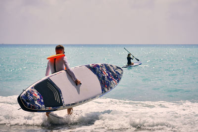 Young man dressed in swimsuit and lifevest carrying sup board towards sea