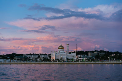 Mosque by river in city against sky during sunset