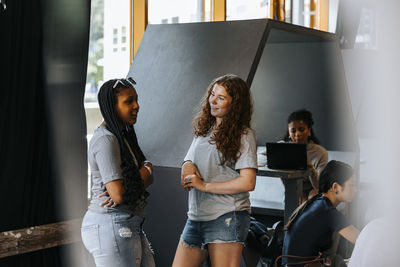Female friends talking with each other while standing near hexagon structure in cafeteria at junior high school