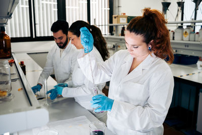 Concentrated female scientist in medical uniform standing near colleagues while pouring colorful chemical liquid from pipette into glass flask working in modern light laboratory