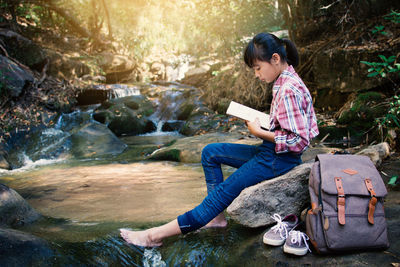 Girl reading book while sitting on rock at river