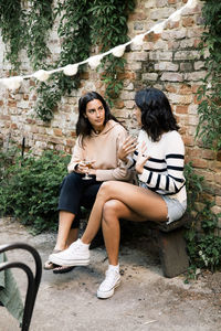 Female friends talking to each other while sitting on bench near brick wall at garden party