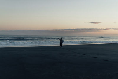 Mid distance view of woman carrying surfboard while walking at beach during sunset