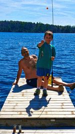 Portrait of shirtless father with son on jetty over lake against sky during sunny day