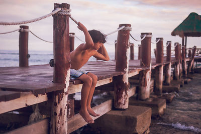Full length of shirtless boy on wooden post against sky