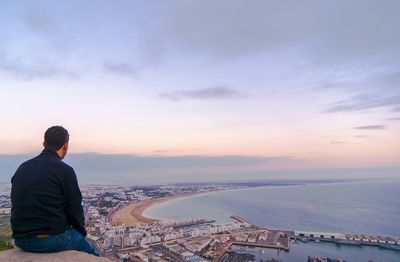 Rear view of man sitting on rock looking at view against sky during sunset