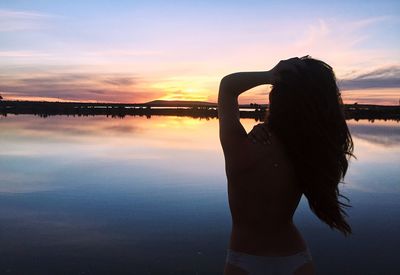 Rear view of topless woman standing at beach during sunset