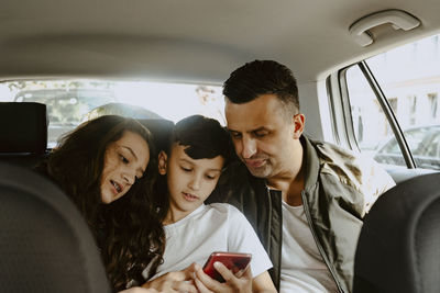 Boy using mobile phone sitting with father and sister in electric car