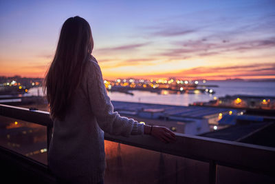 Rear view of woman looking at cityscape against sky during sunset