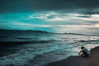 Man drawing while crouching at beach against sky