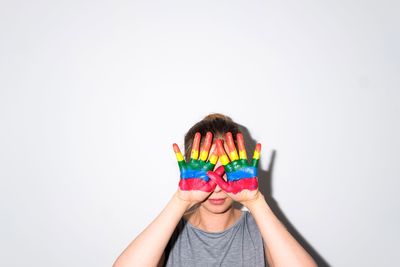 Young woman with colorful painted hands covering face while standing against wall
